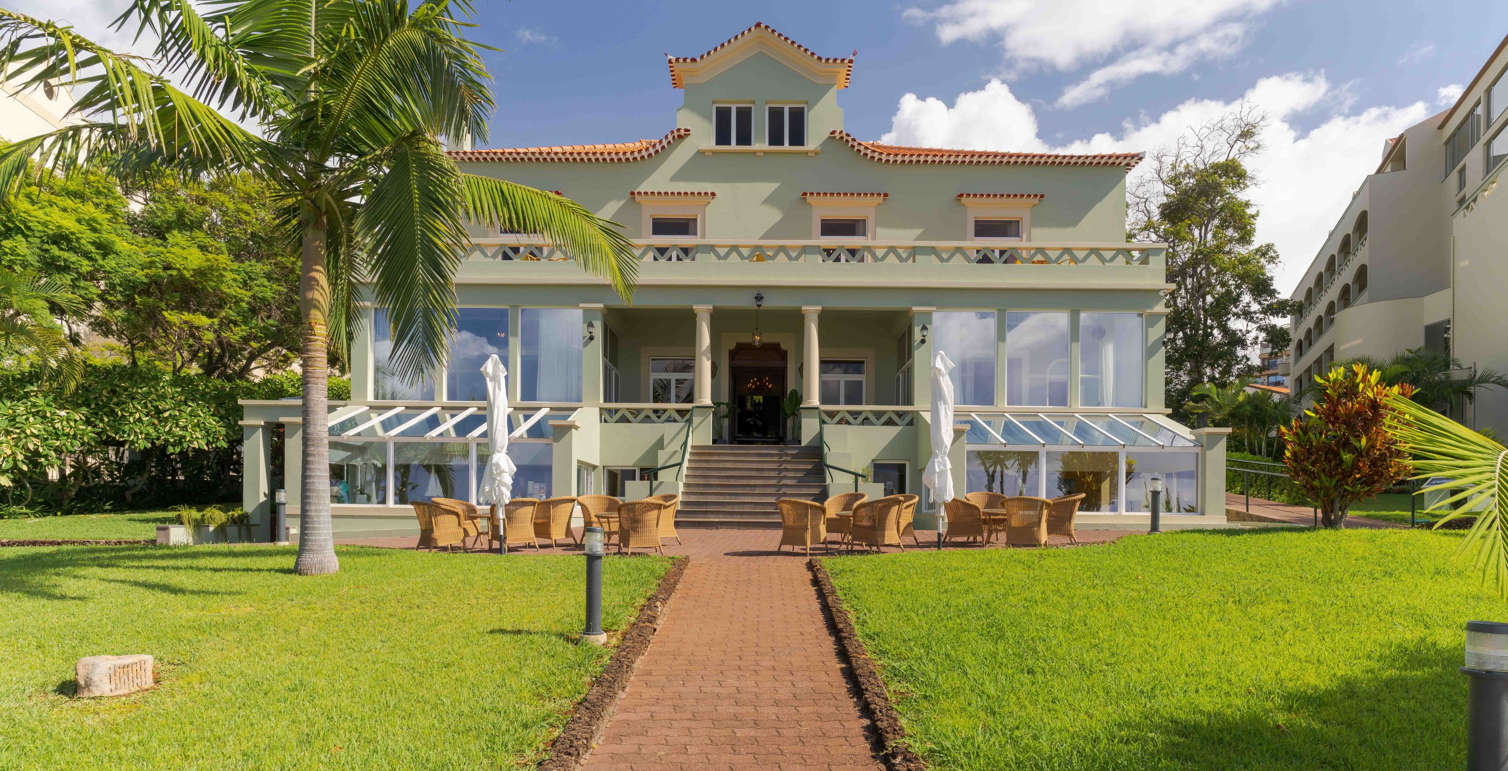 Edificio verde de la recepción del Pestana Vila Lido Madeira, rodeado de césped verde y mesas y sillas en el exterior