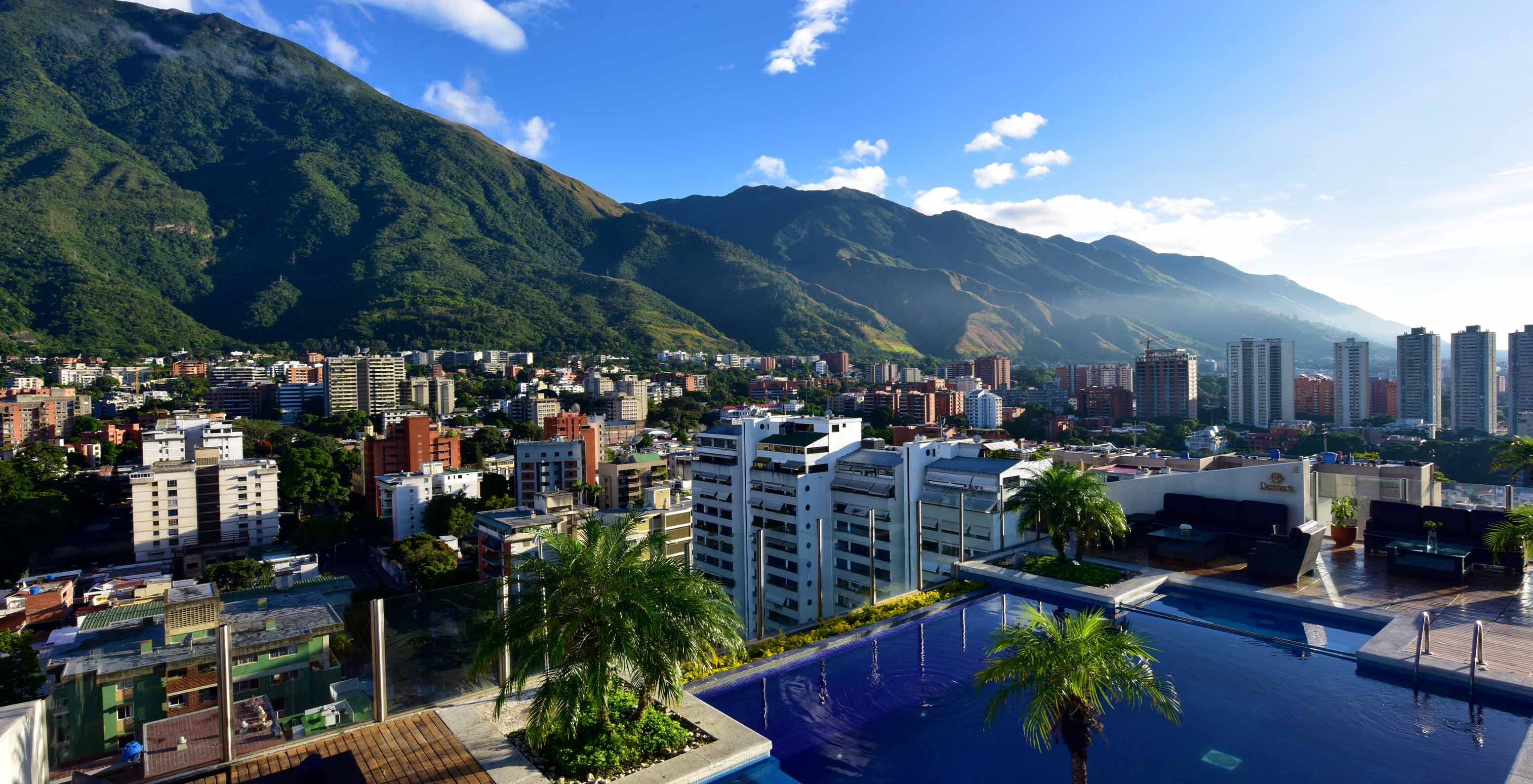 Vista aérea de la piscina del Pestana Caracas, con palmeras y vista a altos edificios y montañas