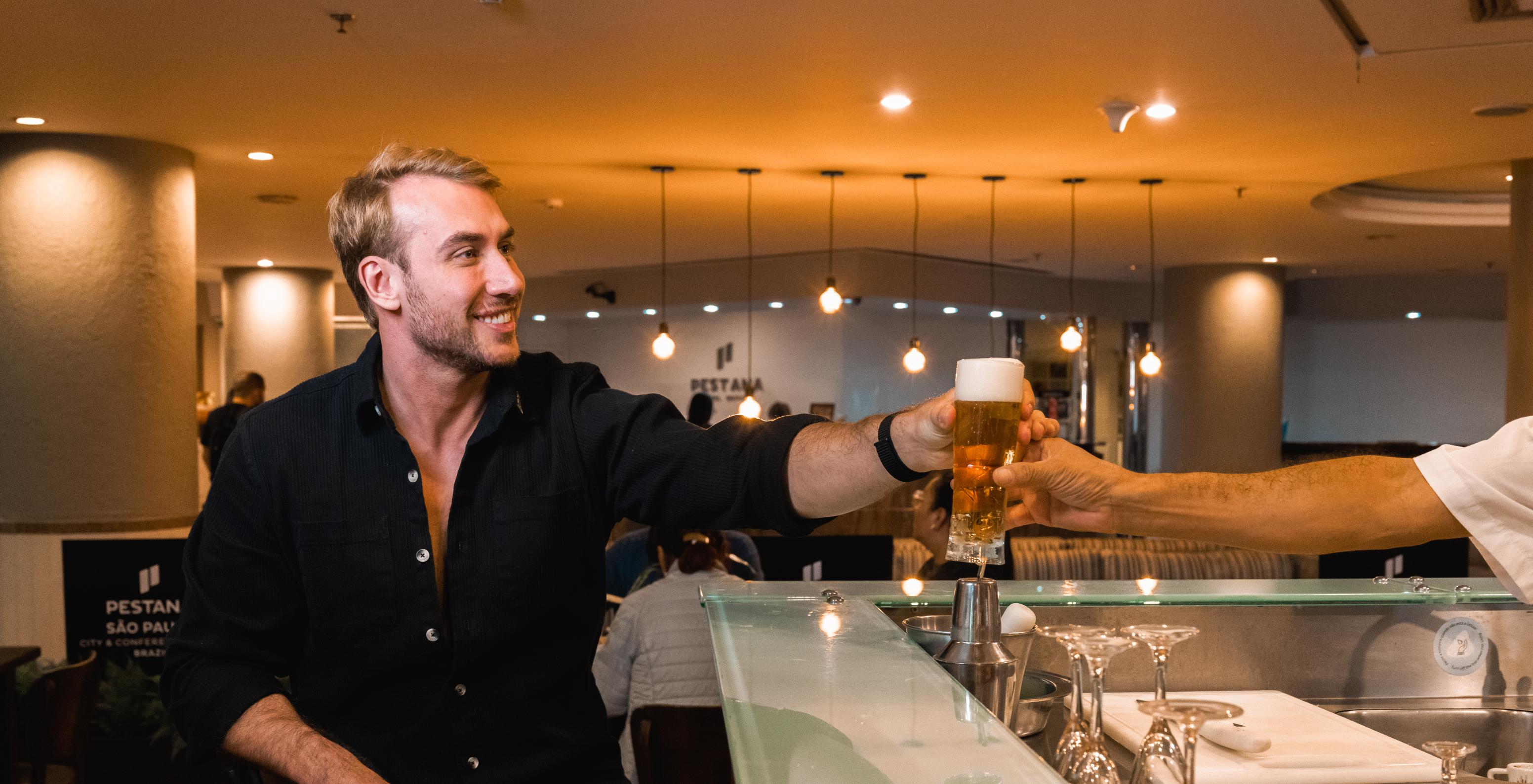 Chico sonriente sentado en banco alto es servido de una cerveza en el Lobby bar de hotel 4 estrellas en São Paulo