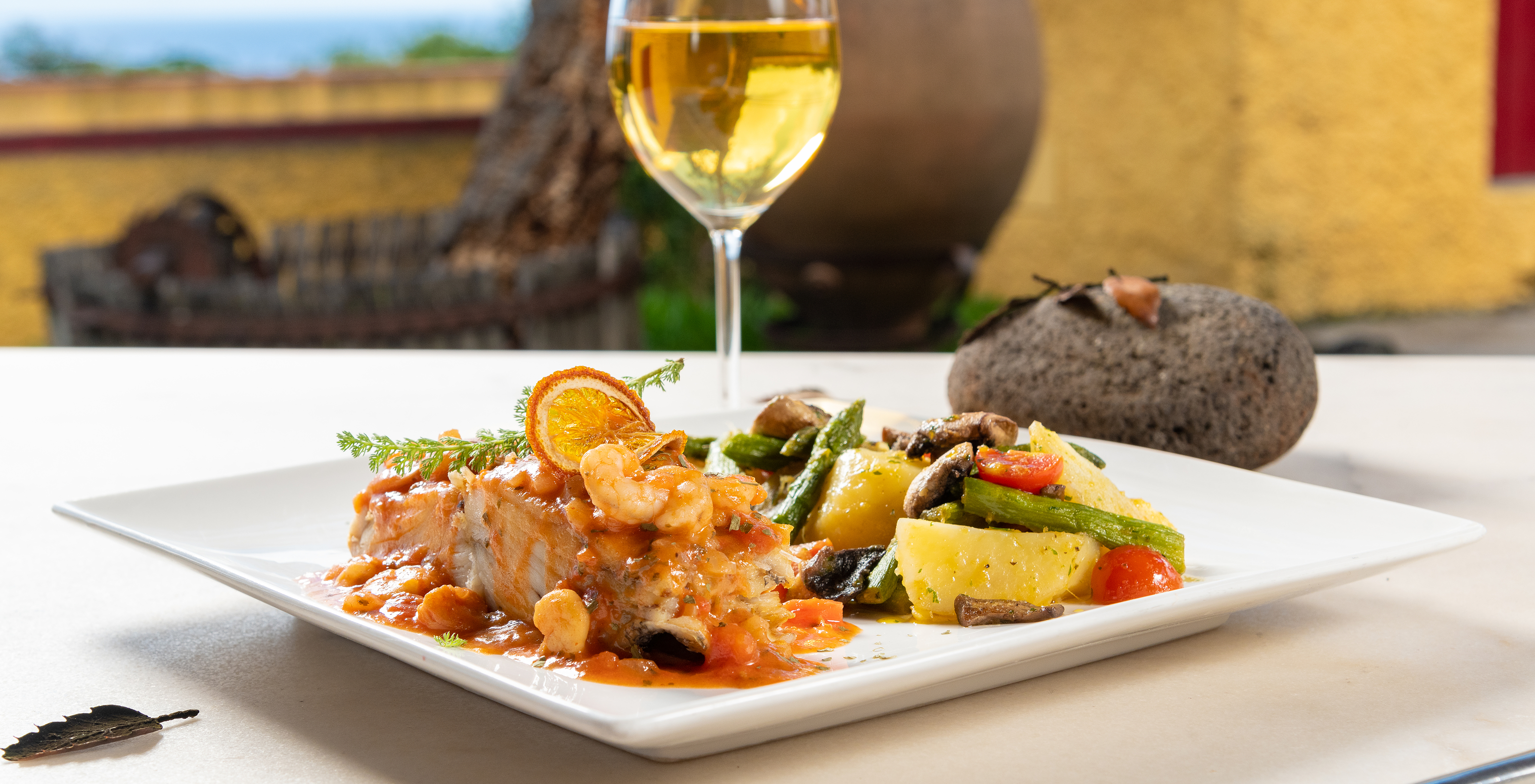 Plato de filete de pescado con camarones, naranja, patata, tomate y otras verduras, con una copa de vino y vista al mar