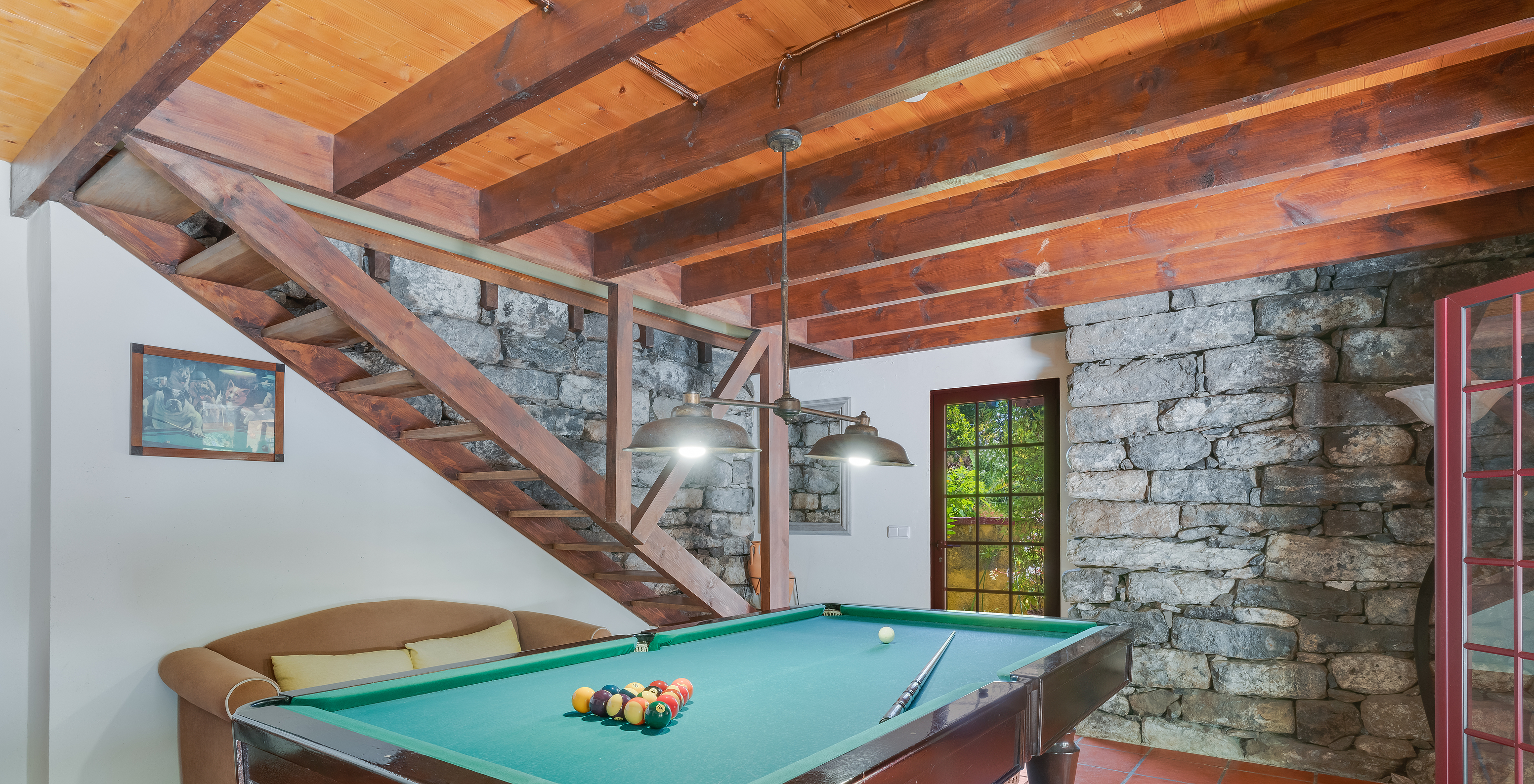 Sala de juegos con mesa de billar, sofá, escaleras de madera y paredes de piedra, con puertas y ventanas al exterior