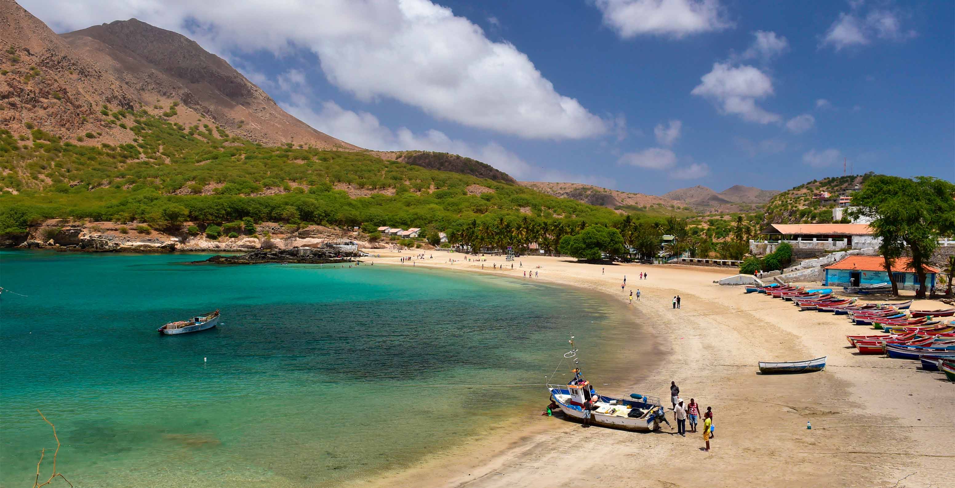 Vue sur une plage avec eau bleu clair, sable blanc, bateaux de pêche et une montagne en arrière-plan