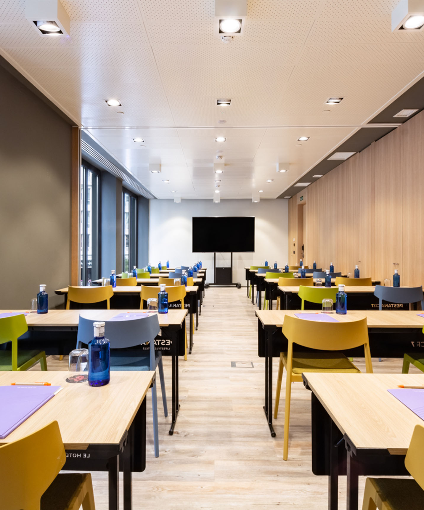 Salle d'événements et de réunions avec tables en bois avec chaises jaunes, vertes et bleues, avec télévision et murs en bois