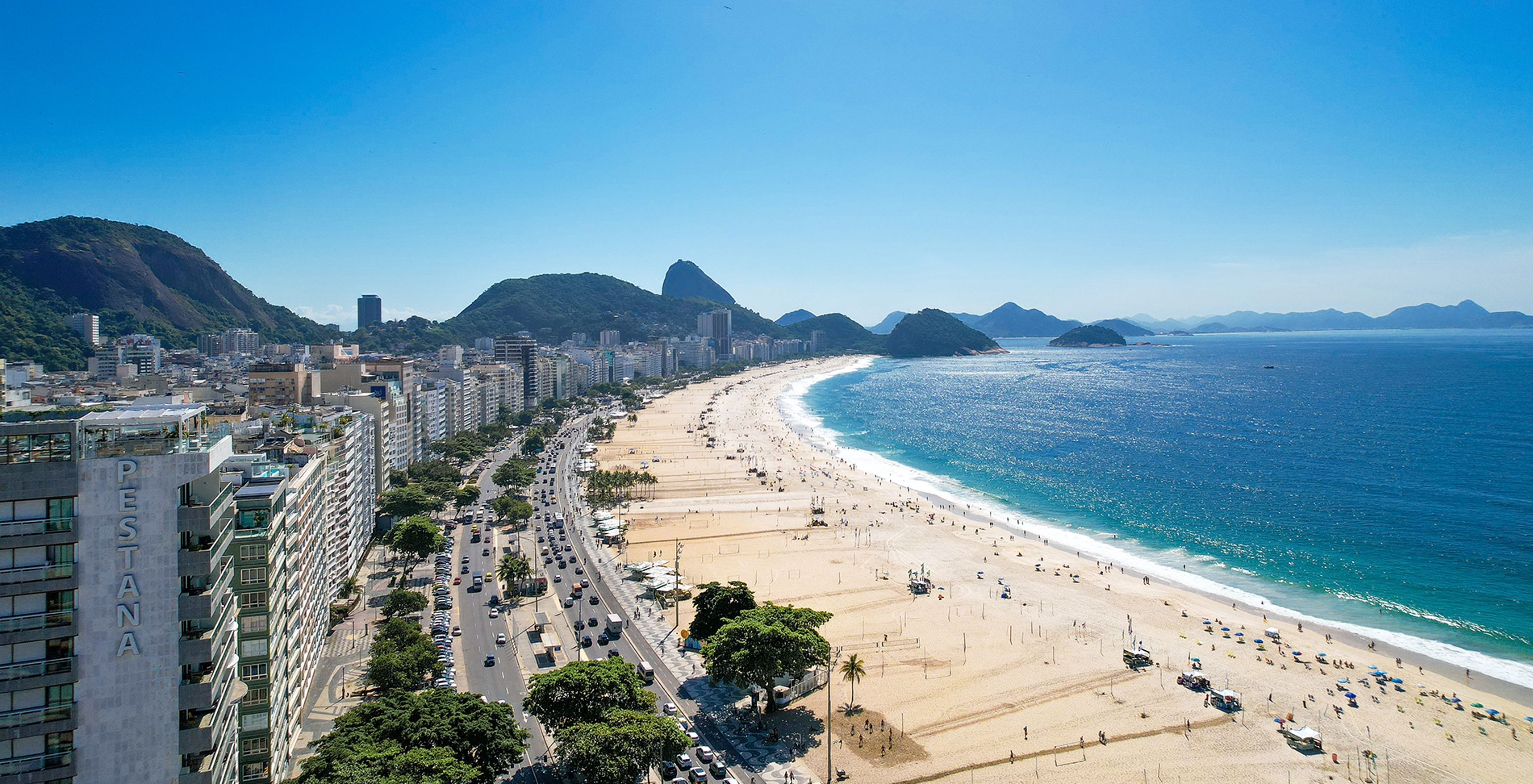 Strand van Copacabana met blauwe zee en bergen, uitgestrekt goudkleurig zand en een trottoir met zwart-witte golvende tegels