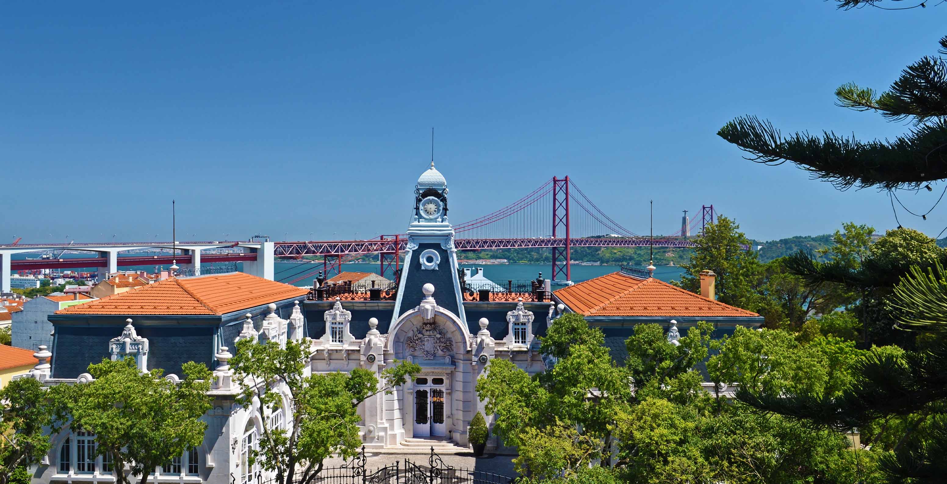 Uitzicht vanuit Pestana Palace Lisboa op de 25 de Abril-brug en de Taag, met verschillende gebouwen en bomen