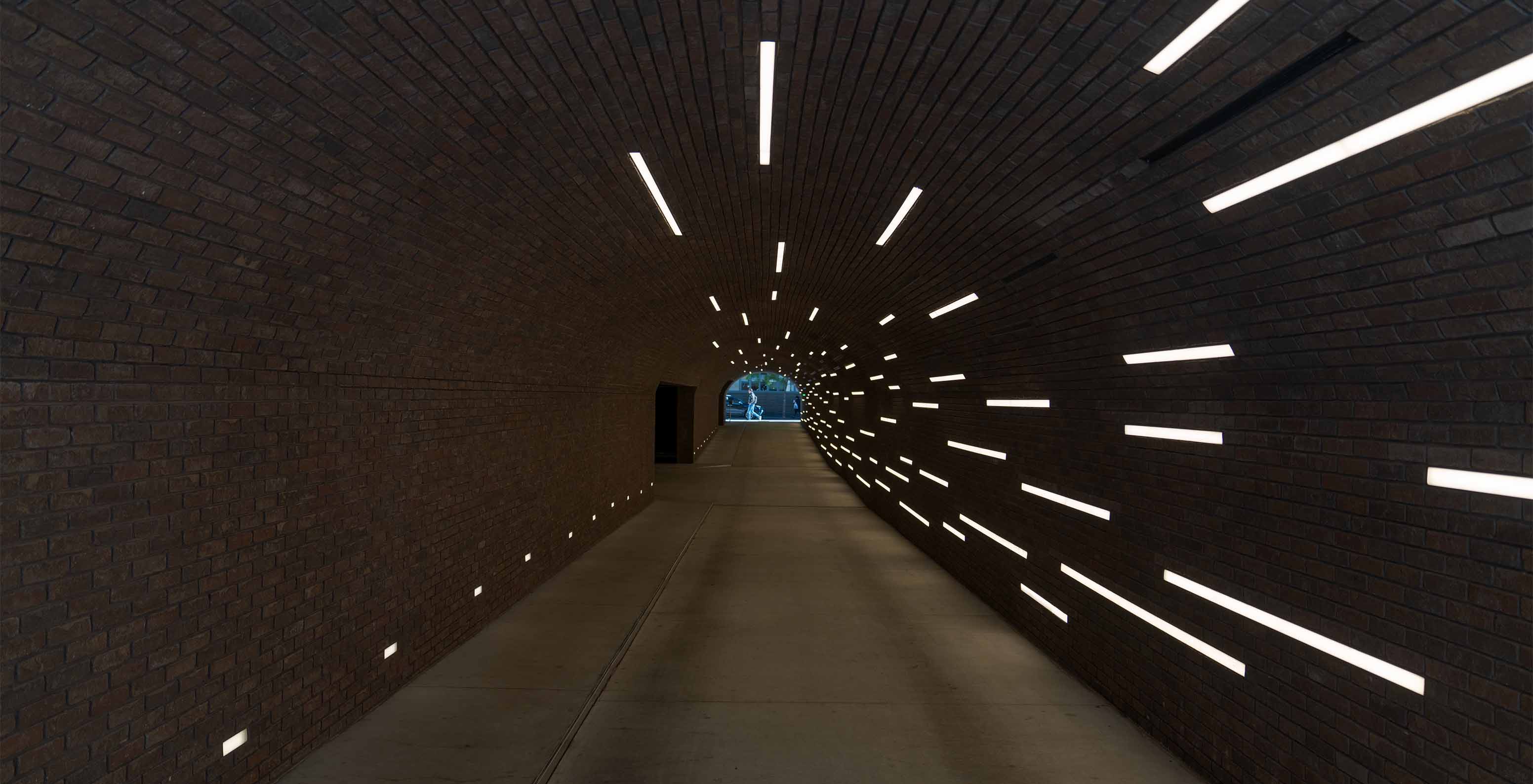 Zicht van binnenin een tunnel met betonnen vloer en bakstenen muren, verlicht door fluorescerende lichten