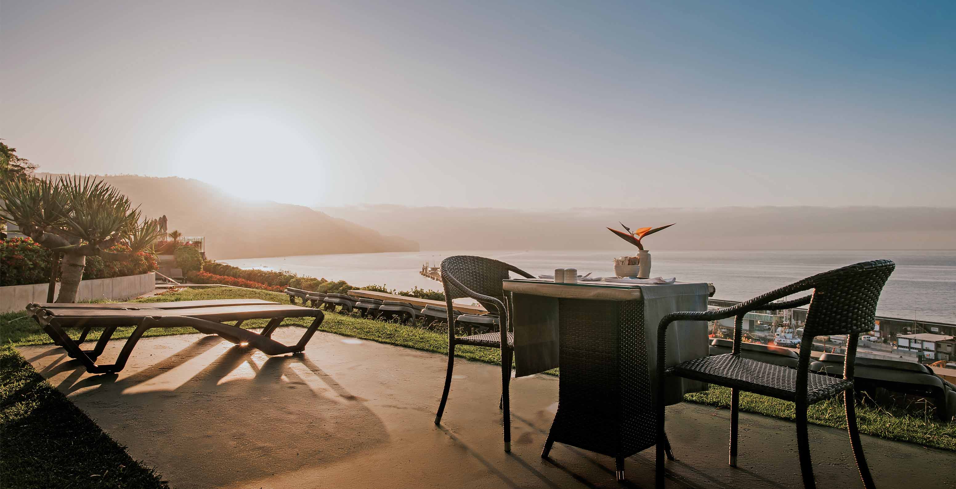 Vista para o pôr do sol, de uma esplanada com uma mesa e espreguiçadeiras e com vista para o mar, no Pestana Casino Park