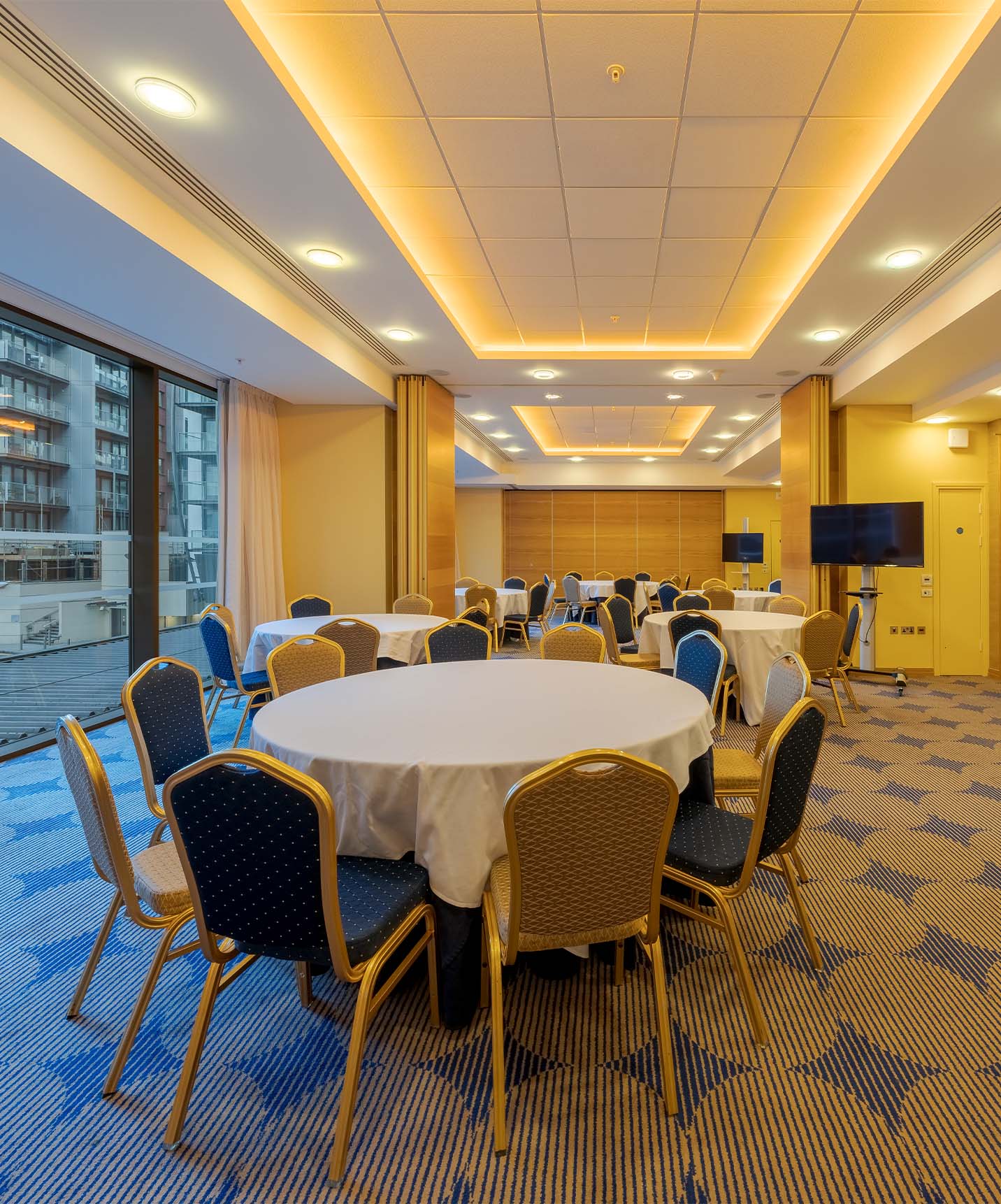 Sala de reuniões com mesas redondas, divisórias em madeira, televisões e janelas grandes com vista cidade