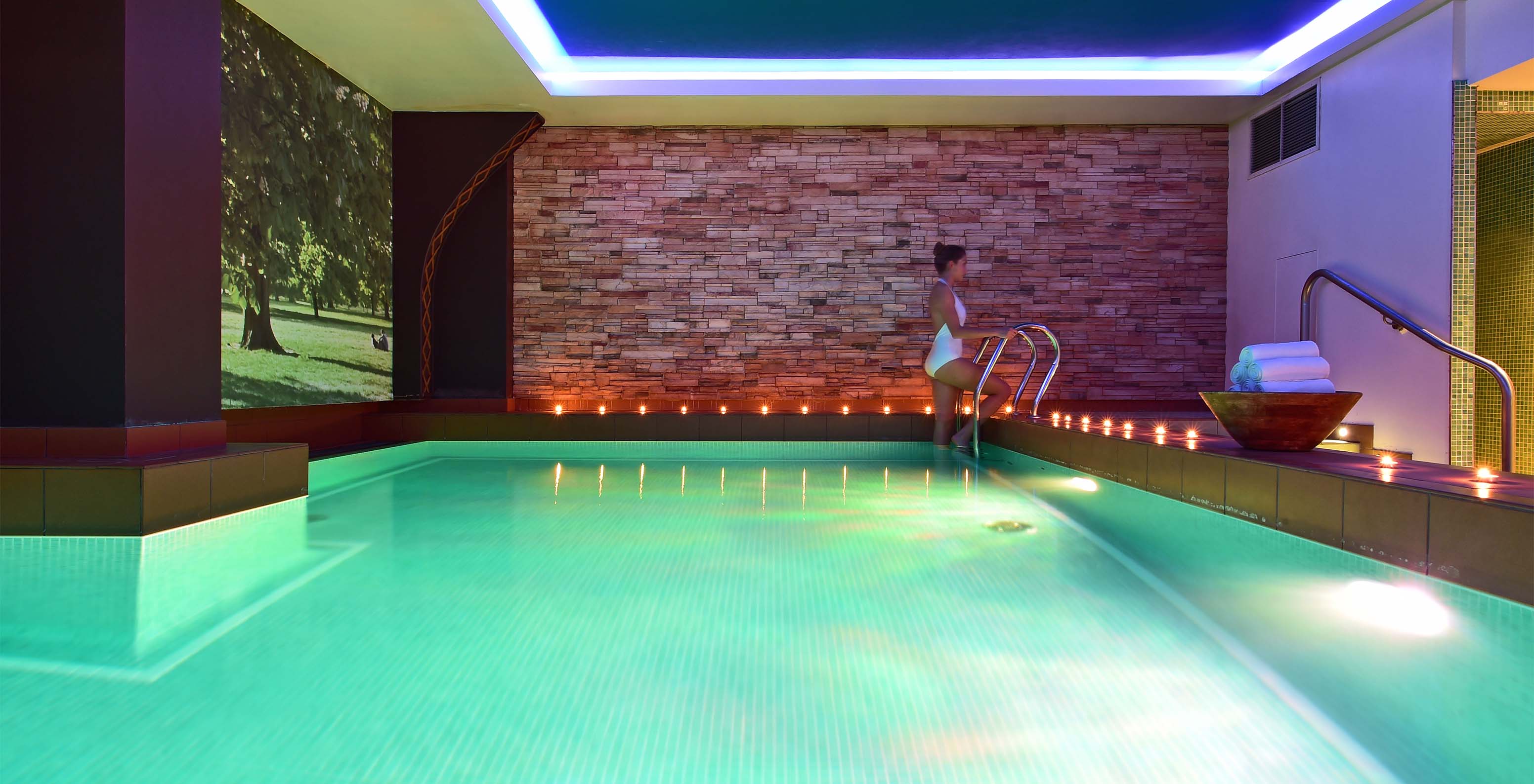 Piscina interior com iluminação interior azul e exterior laranja, com uma senhora a subir as escadas e luzes azuis no teto