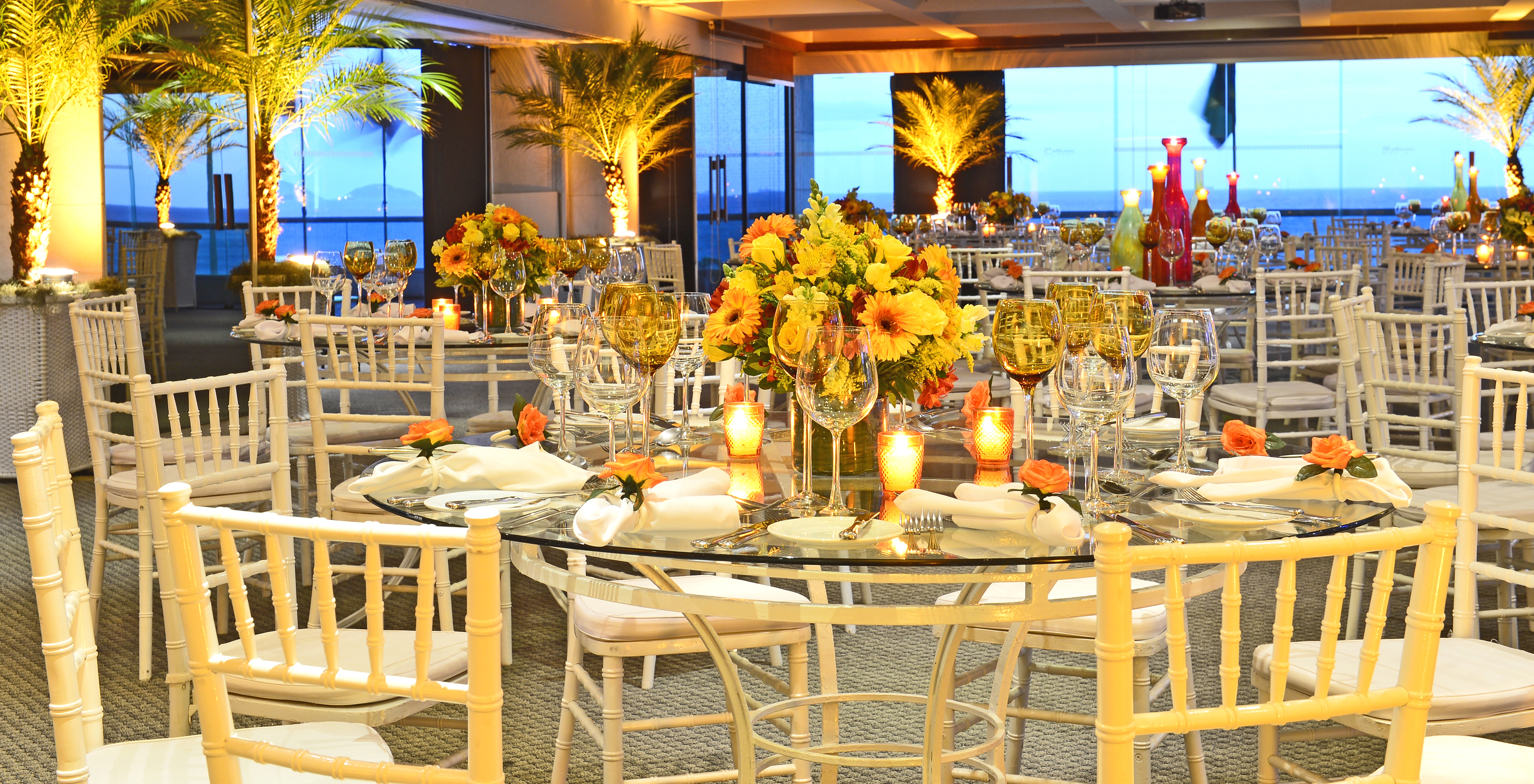 Sala de eventos do Hotel Junto à Praia de Copacabana, com palmeiras iluminadas e várias mesas com centros de flores