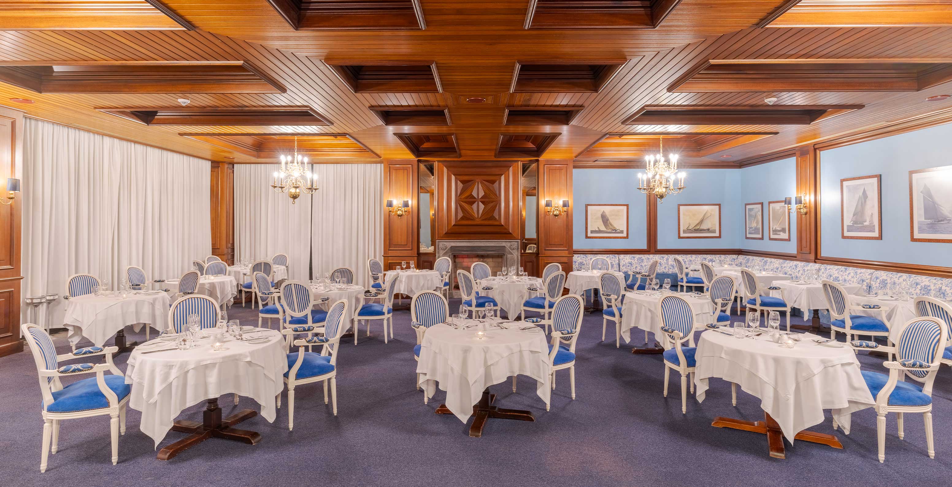 Parte interior do restaurante do Pestana Bahia Praia, com mesas de toalha branca, cadeiras azuis às riscas e teto de madeira